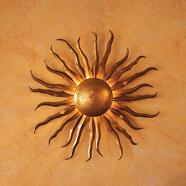 Eine goldfarbene, sonnenförmige Leuchte, die an einer strukturierten Wand in einem warmen Orangeton angebracht ist. Die Leuchte besteht aus einer zentralen Kugel, von der wellenförmige Metallstrahlen ausgehen, die einen dezenten, warmen Schein verbreiten.