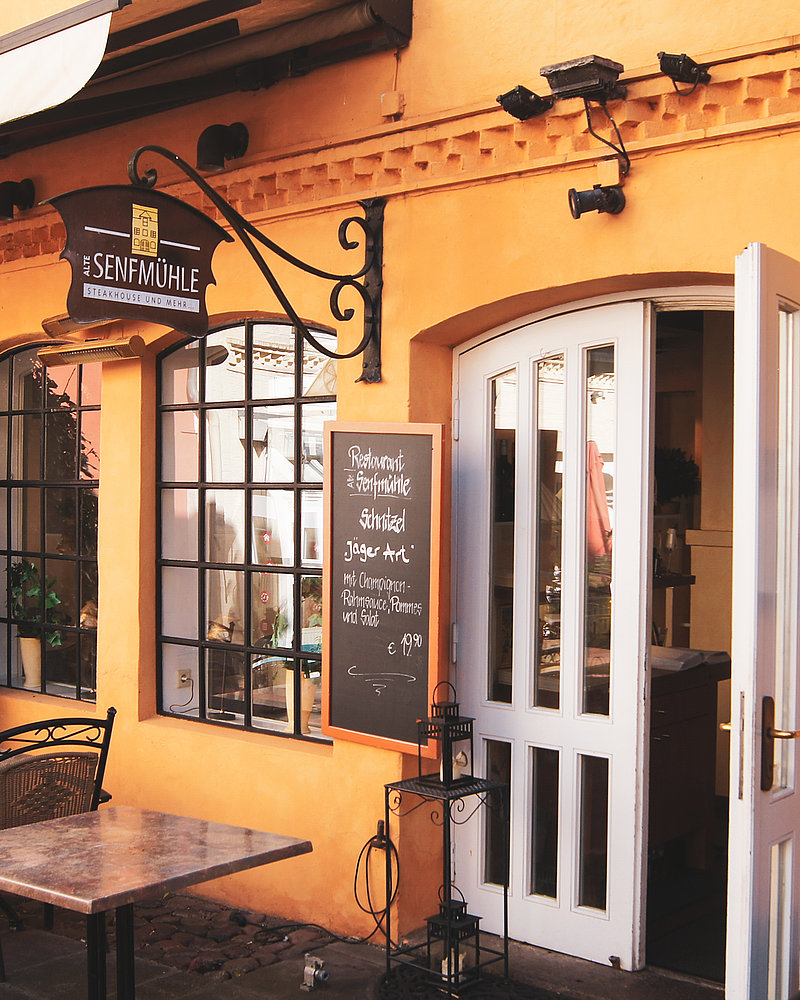 Ein Restaurant mit orangefarbenen Wänden namens „Alte Senfmühle“ mit einer offenen weißen Tür, einer Speisekarte auf einer Kreidetafel, auf der „Schnitzel nach Jägerart“ für 19,90 € angeboten wird, und Sitzgelegenheiten im Freien mit Metallstühlen und -tischen auf einer Terrasse mit Kopfsteinpflaster.