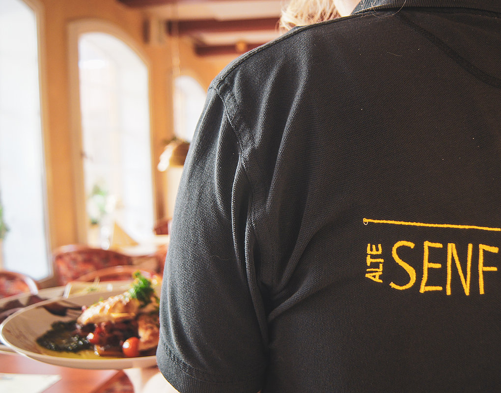 Eine Person in einem schwarzen Hemd, auf dessen Rücken „Alte Senf Mühle“ gestickt ist, serviert in einem gemütlich beleuchteten Restaurant einen Teller mit Essen.