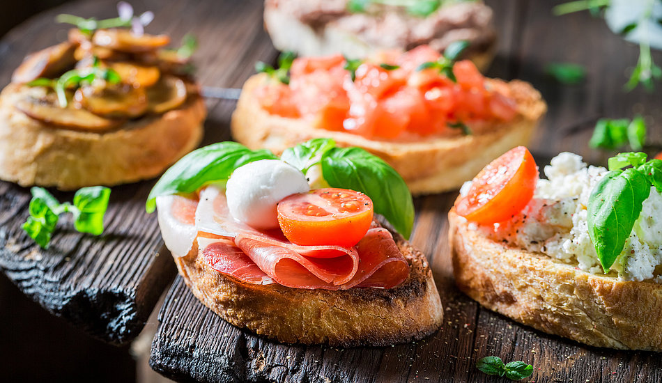 Verschiedene Bruschetta-Variationen mit Belägen wie Mozzarella, Tomaten, Basilikum, Wurstwaren, Pilzen, Ricotta und Kräutern auf rustikalem geröstetem Brot, serviert auf einem Holzbrett.