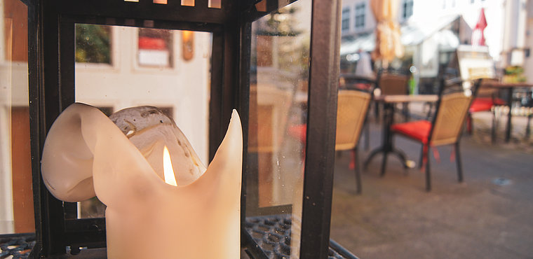 Eine Nahaufnahme einer schmelzenden Kerze in einer schwarzen Metalllaterne, deren kleine Flamme zu sehen ist. Der Hintergrund ist leicht unscharf und zeigt Stühle und Tische auf der sonnigen Terrasse eines Straßencafés.