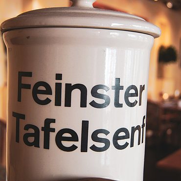 Im Mittelpunkt steht ein großer Keramikbehälter mit der Aufschrift „Feinster Tafelsenf“ in fetten schwarzen Buchstaben, während im Hintergrund der leicht unscharf dargestellte Speisesaal des Restaurants und Holztische zu sehen sind.