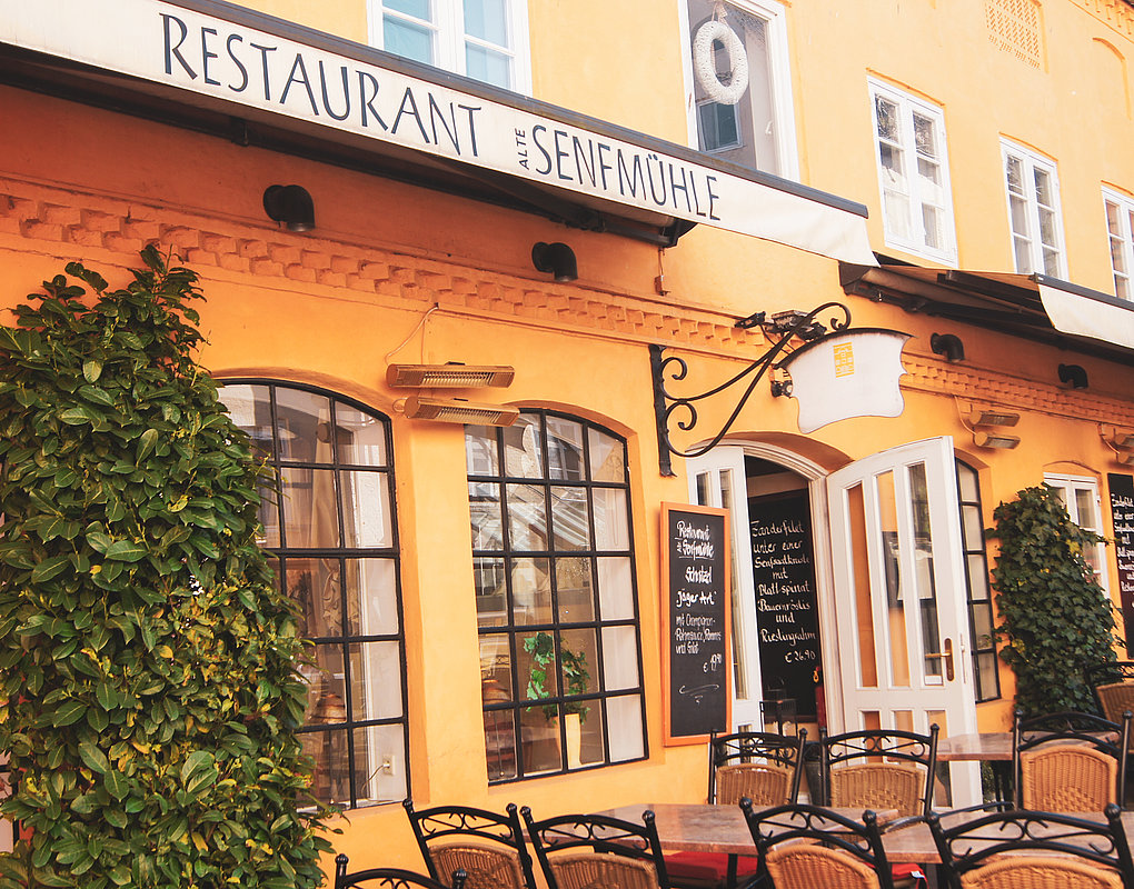 Die Außenterrasse des Restaurants „Alte Senfmühle“, eines Gebäudes mit orangefarbenen Wänden, großen Bogenfenstern, Kletterpflanzen und Metallstühlen und -tischen, die zum Essen aufgestellt sind. Neben der offenen weißen Tür steht eine Speisekarte.