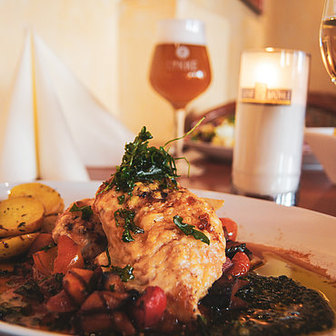 Ein Teller mit Bratkartoffeln und Hähnchen, garniert mit Blattgemüse, umgeben von buntem Gemüse und Soße. Im Hintergrund stehen auf einem Holztisch ein Glas Weißwein, ein Bier und eine brennende Kerze.