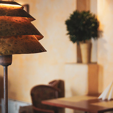 Ein warmes, sanft beleuchtetes Restaurantinterieur mit einem einzigartigen Lampenschirm aus Messing im Vordergrund, Topfpflanzen, Tischen und Stühlen, die im Hintergrund verschwommen wirken.