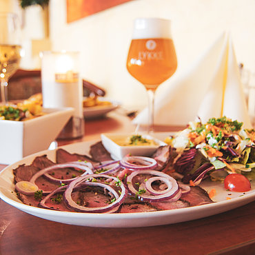 Ein Teller mit dünn geschnittenem Roastbeef, garniert mit roten Zwiebelringen, serviert mit einem frischen Beilagensalat und einer kleinen Schale Soße, auf einem Holztisch mit einem Glas Bier, einem Weinglas und einer Serviette in einem Restaurant.