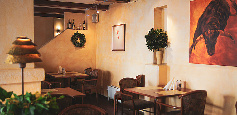 Ein gemütliches Restaurant mit hellgelben Wänden, Holztischen und -stühlen, Topfpflanzen, Weinflaschen auf einem Sims, Servietten auf den Tischen und einem großen Gemälde eines Stiers an der Wand. Die warme Beleuchtung sorgt für eine einladende Atmosphäre.