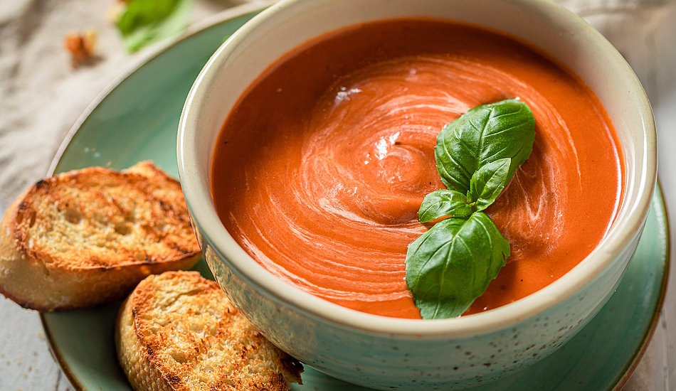 Eine Schüssel cremige Tomatensuppe, garniert mit frischen Basilikumblättern, serviert auf einem grünen Teller mit gerösteten Brotscheiben als Beilage.